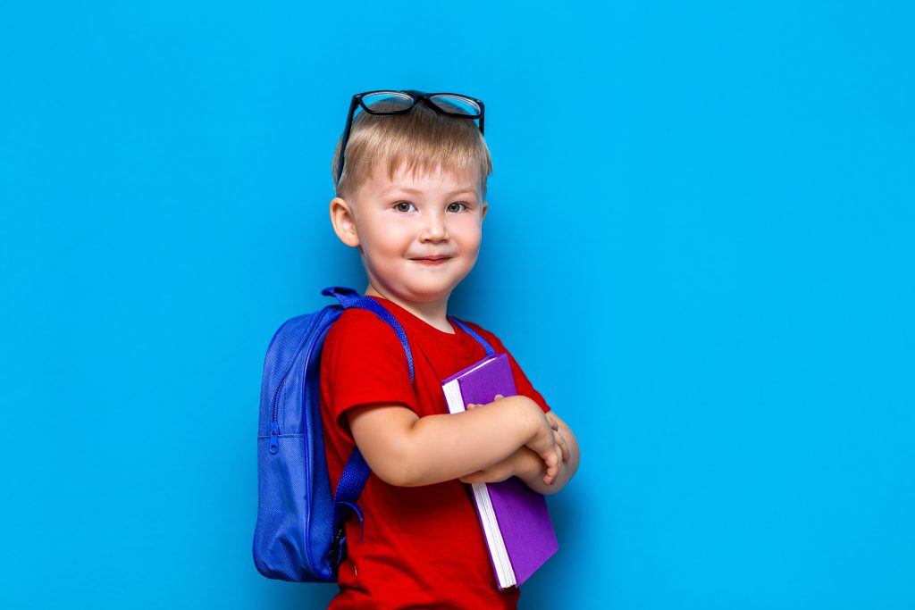 small happy smiling boy with glasses on his head, book in hands, schoolbag on his shoulders. back to school. ready to school